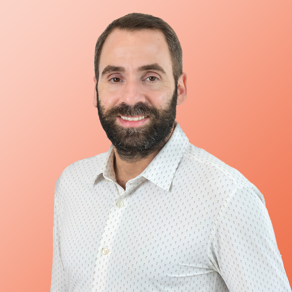 Un homme avec une barbe et une moustache, vêtu d'une chemise blanche à motifs boutonnée d'AMO marché public, sourit à la caméra sur un fond couleur pêche.